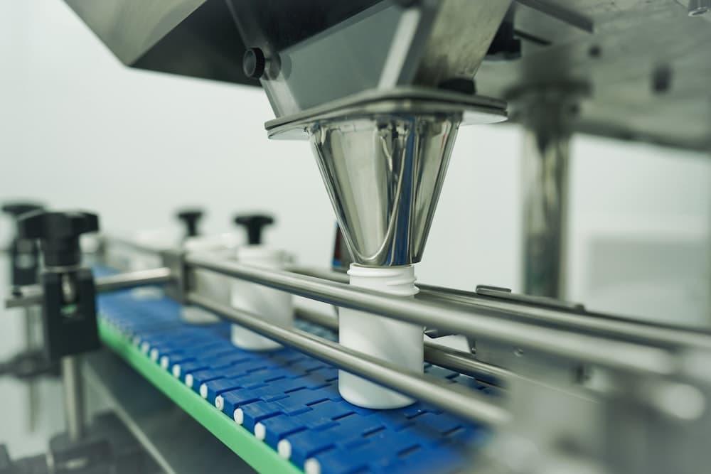 A filling head dosing white bottles on a conveyor, unlabelled bottles under a rotary filler, amber jars with blank labels, an open amber bottle with capsules beside it, rows of plain ceramic cups, and a container terminal from the air